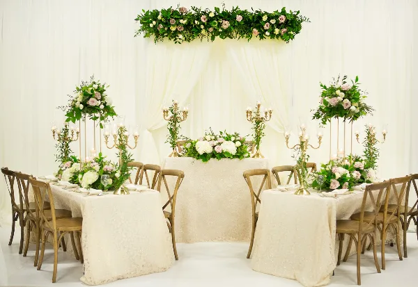 A wedding head table setup featuring white linens, wooden chairs, tall floral centerpieces, and a floral backdrop against white drapery.