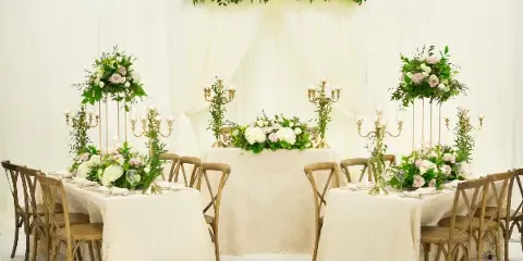 A wedding head table setup featuring white linens, wooden chairs, tall floral centerpieces, and a floral backdrop against white drapery.