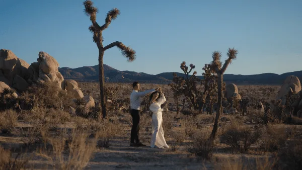 A cinematic wide shot of a bride and groom dancing in a desert landscape with Joshua trees and mountains in the background.