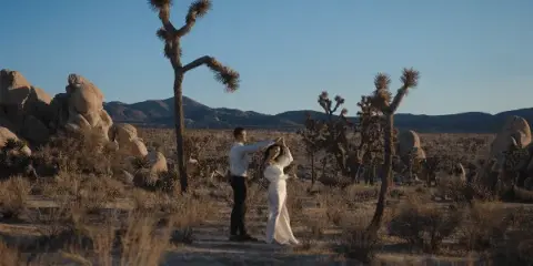 A cinematic wide shot of a bride and groom dancing in a desert landscape with Joshua trees and mountains in the background.
