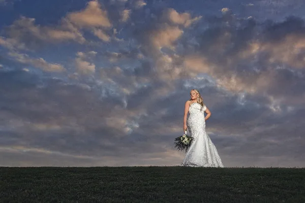 A bride in a white wedding dress holding a bouquet stands in a grassy field under a dramatic cloudy sky.