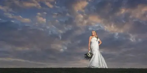 A bride in a white wedding dress holding a bouquet stands in a grassy field under a dramatic cloudy sky.