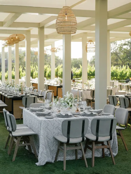 Elegant outdoor wedding reception table setting with white floral arrangements, textured table linens, and woven pendant lighting.