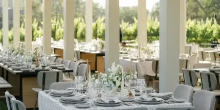 Elegant outdoor wedding reception table setting with white floral arrangements, textured table linens, and woven pendant lighting.