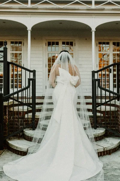 A bride in a white wedding dress and veil stands on the stone steps of a white building with arched windows.