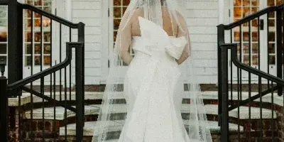 A bride in a white wedding dress and veil stands on the stone steps of a white building with arched windows.