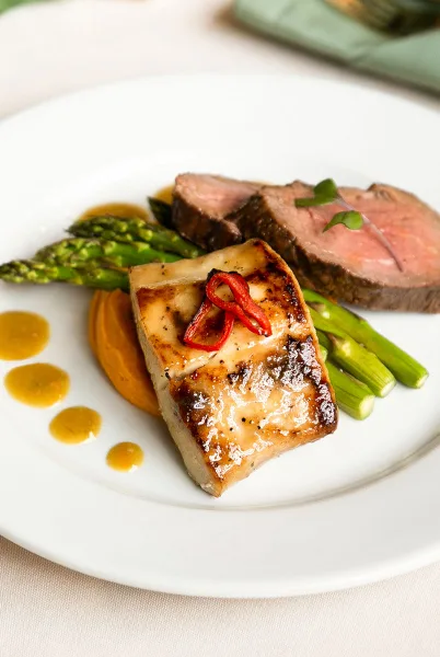 A plated catering entree featuring seared fish, sliced steak, and asparagus on a white plate.