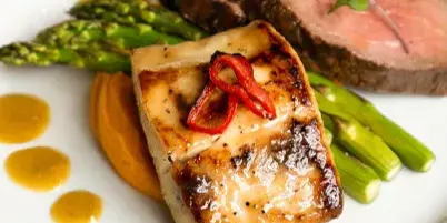 A plated catering entree featuring seared fish, sliced steak, and asparagus on a white plate.