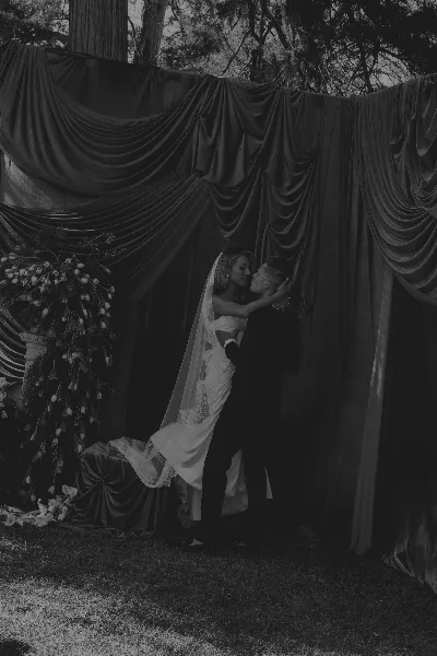 A black and white photograph of a bride and groom kissing in front of a draped fabric backdrop with floral arrangements.