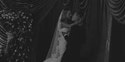 A black and white photograph of a bride and groom kissing in front of a draped fabric backdrop with floral arrangements.
