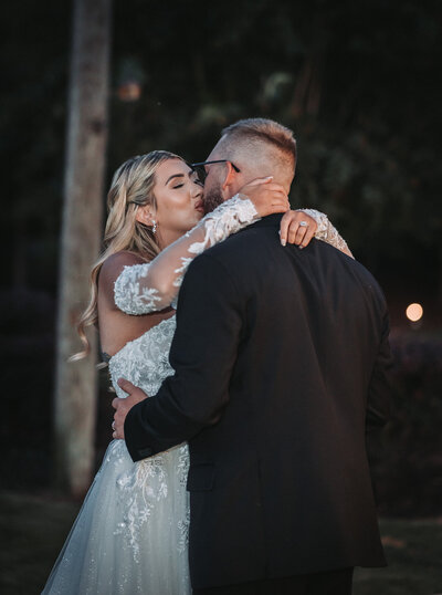 A bride in a white lace gown and a groom in a black suit embracing and kissing outdoors at dusk.