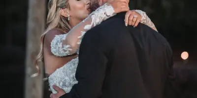 A bride in a white lace gown and a groom in a black suit embracing and kissing outdoors at dusk.