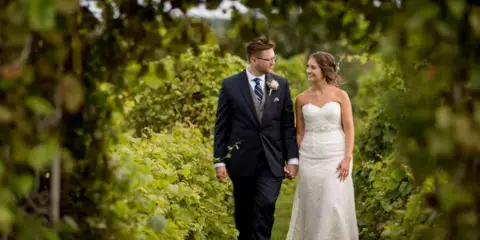 A bride and groom walking hand-in-hand through a lush green vineyard.