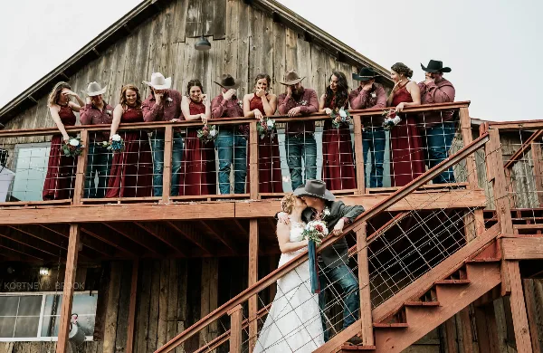 A wedding party posing on the balcony of a rustic wooden barn with a couple kissing on the stairs below.