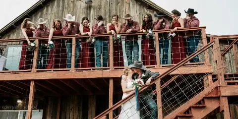 A wedding party posing on the balcony of a rustic wooden barn with a couple kissing on the stairs below.