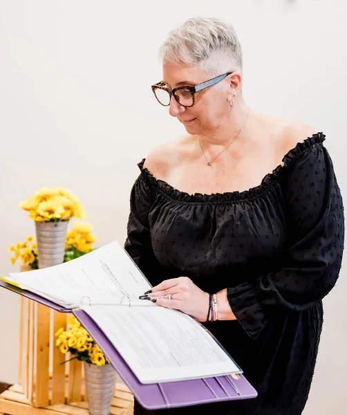 A woman with short grey hair and glasses, wearing a black off-the-shoulder top, stands reading from a purple binder containing a wedding script.