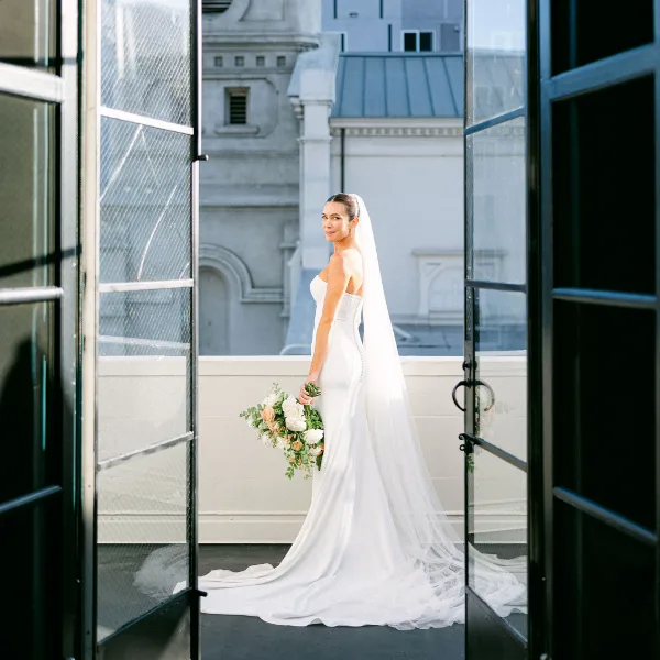 A bride in a white wedding dress and long veil stands on a balcony holding a bouquet, looking over her shoulder.