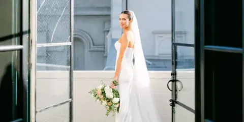 A bride in a white wedding dress and long veil stands on a balcony holding a bouquet, looking over her shoulder.