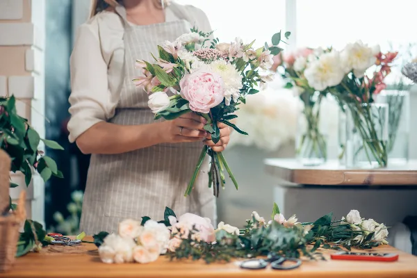 Florist in an apron holding a freshly arranged bouquet of pink and white flowers at a workbench with loose stems and scissors.
