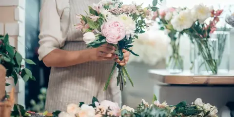 Florist in an apron holding a freshly arranged bouquet of pink and white flowers at a workbench with loose stems and scissors.