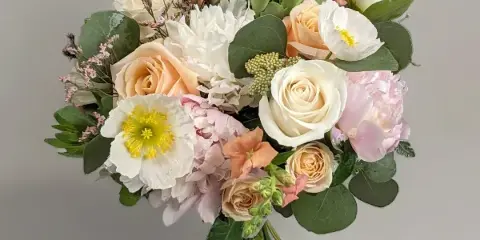 A hand-tied floral bouquet containing white and peach roses, ranunculus, and greenery against a neutral background.