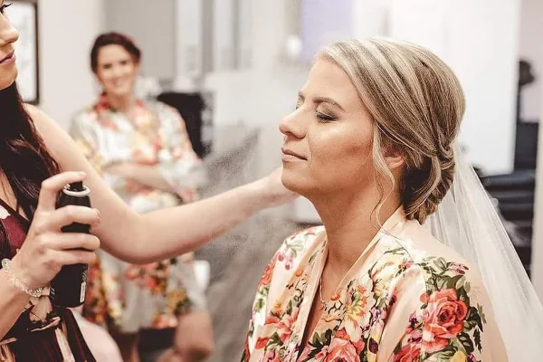 A makeup artist using an airbrush tool to apply makeup to a bride wearing a floral robe and veil.