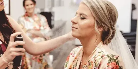 A makeup artist using an airbrush tool to apply makeup to a bride wearing a floral robe and veil.
