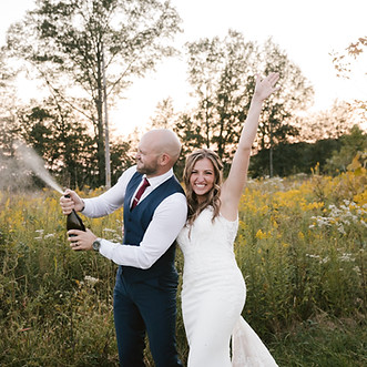 A bride and groom celebrating outdoors in a field, with the groom popping a bottle of champagne.