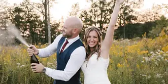 A bride and groom celebrating outdoors in a field, with the groom popping a bottle of champagne.