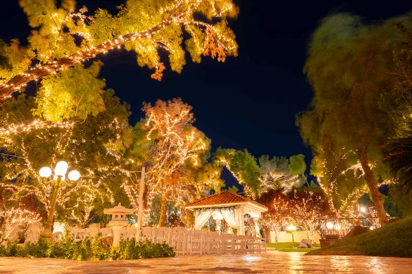 Nighttime view of an outdoor wedding venue featuring a gazebo, white chairs, and trees illuminated with string lights.