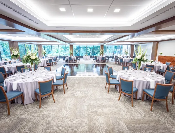 A large, well-lit event ballroom set up for a wedding reception with round tables, blue chairs, and floral arrangements.