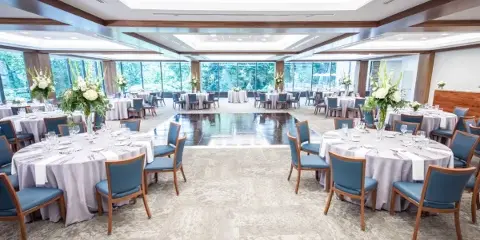 A large, well-lit event ballroom set up for a wedding reception with round tables, blue chairs, and floral arrangements.