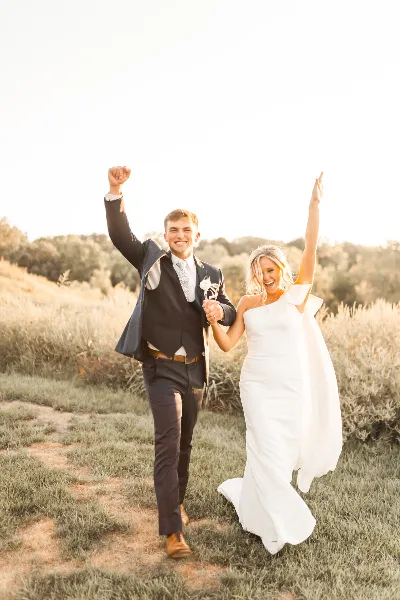 A bride and groom walking through a grassy field with arms raised in celebration during golden hour.