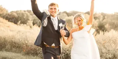 A bride and groom walking through a grassy field with arms raised in celebration during golden hour.