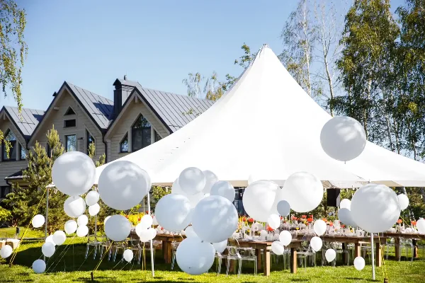 An outdoor event setup featuring a large white peak tent, long wooden tables, and clear chairs on a lawn, decorated with white balloons.