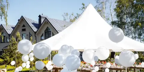 An outdoor event setup featuring a large white peak tent, long wooden tables, and clear chairs on a lawn, decorated with white balloons.