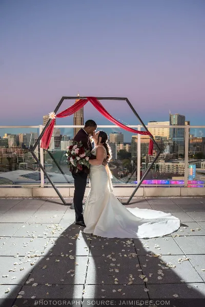 A bride and groom sharing a kiss on a rooftop terrace at dusk, framed by a geometric arch with red fabric, with a city skyline in the background.