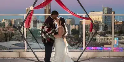 A bride and groom sharing a kiss on a rooftop terrace at dusk, framed by a geometric arch with red fabric, with a city skyline in the background.
