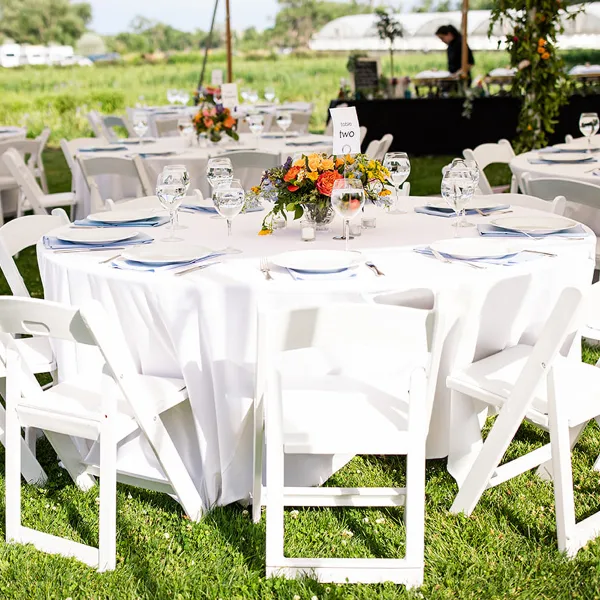 A round banquet table covered in a white tablecloth, set with white plates, blue napkins, and glassware, surrounded by white folding chairs on a grassy lawn.
