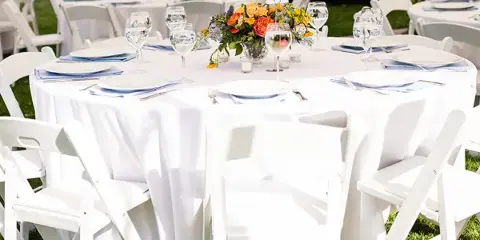 A round banquet table covered in a white tablecloth, set with white plates, blue napkins, and glassware, surrounded by white folding chairs on a grassy lawn.