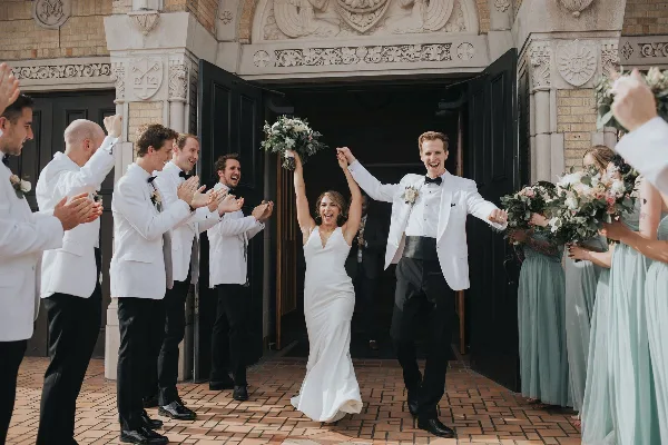 A bride and groom exit a stone building, holding hands and smiling, surrounded by their wedding party.