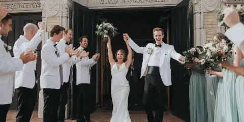 A bride and groom exit a stone building, holding hands and smiling, surrounded by their wedding party.
