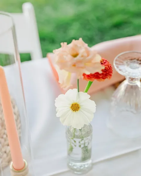 A close-up of a small floral arrangement featuring a peach rose, a red flower, and a white daisy in a small glass vase on a table.