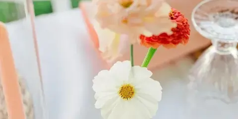 A close-up of a small floral arrangement featuring a peach rose, a red flower, and a white daisy in a small glass vase on a table.