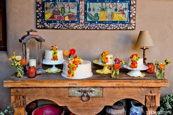 A rustic wooden table displays multiple white wedding cakes decorated with colorful flowers, arranged with candles and floral accents.
