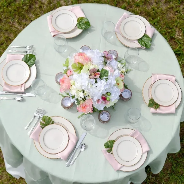 A round table set with a mint green tablecloth, white and gold-rimmed plates, pink napkins, silver flatware, and a large floral centerpiece.