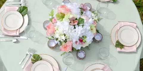 A round table set with a mint green tablecloth, white and gold-rimmed plates, pink napkins, silver flatware, and a large floral centerpiece.