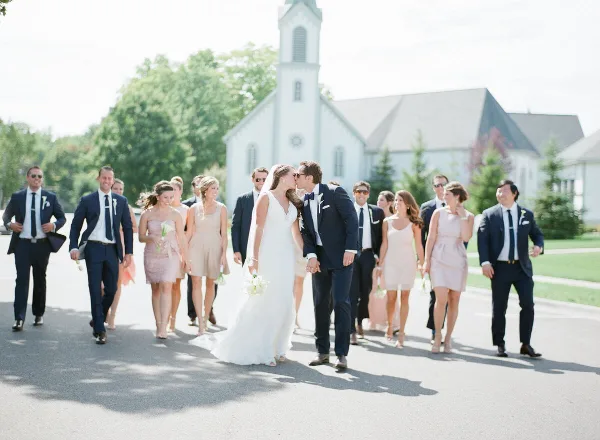 A bride and groom kissing while walking with their wedding party in front of a church.