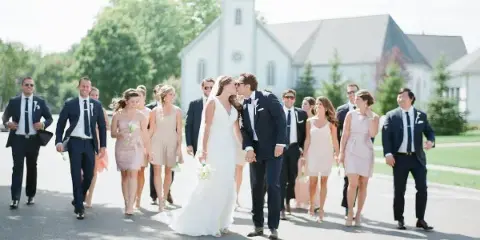 A bride and groom kissing while walking with their wedding party in front of a church.