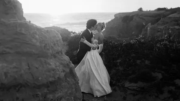 A black and white photograph of a bride and groom kissing on a rocky coastal cliff.